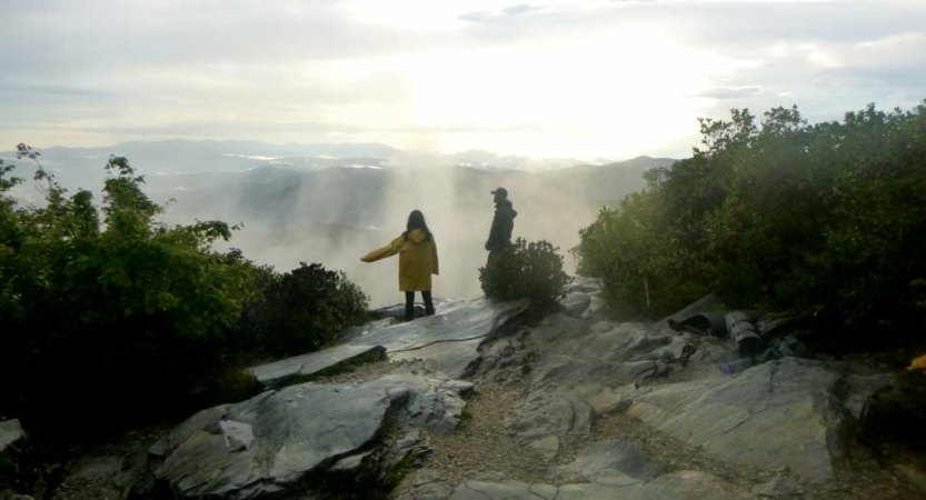 Two people stand on a rock overlook. Beyond them is fog and the outline of the blue ridge mountains. 